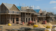 Construction site with partially built brick houses, scaffolding, building materials, and sand piles under a blue sky